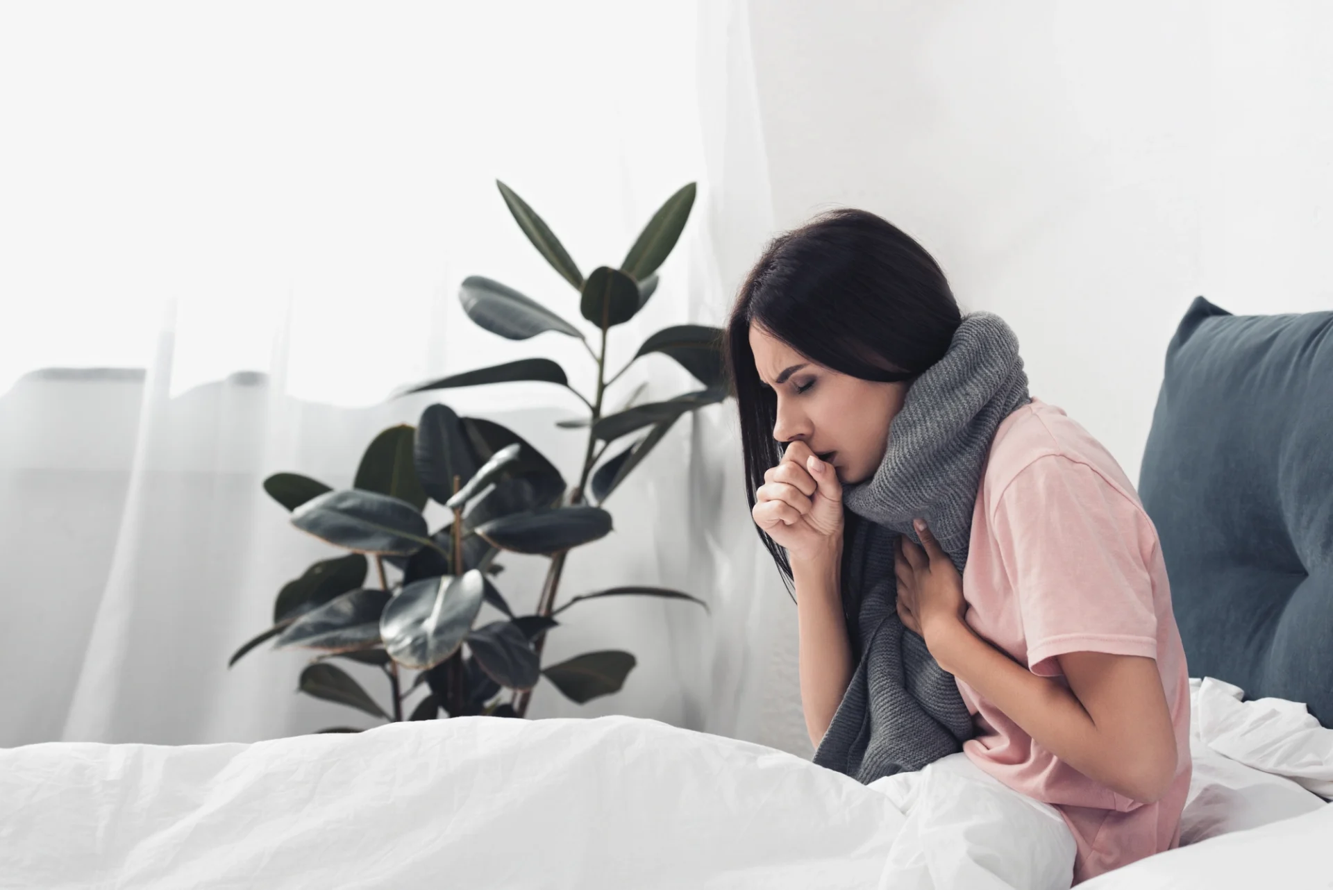 woman sitting up with a scarf on and hand over her mouth as if coughing