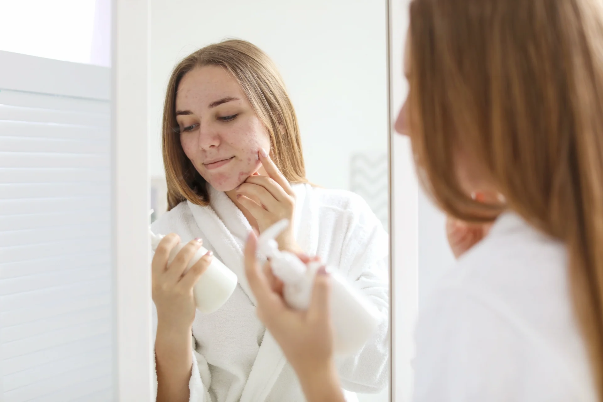 woman with mild acne standing in front of a mirror holding a bottle of face wash